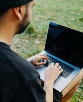 A man sits outdoors on the grass using a laptop, enjoying nature.