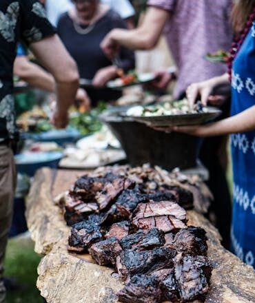 A group of people enjoying a summer barbecue with grilled meat on a wooden table outdoors.