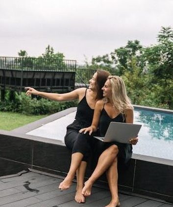 Two women enjoying a serene moment by the pool with a laptop, surrounded by nature.